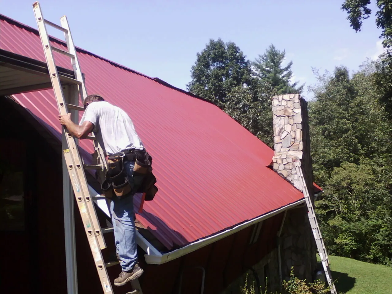 Roofer on a ladder beside a red metal roof and stone chimney — Soffit & Fascia Repair by Roofing Brothers in Ivydale